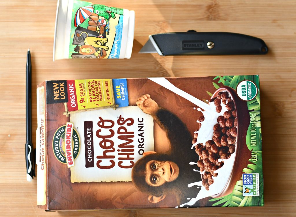 An empty cereal box, cup, and box cutter on top of a cutting board. 