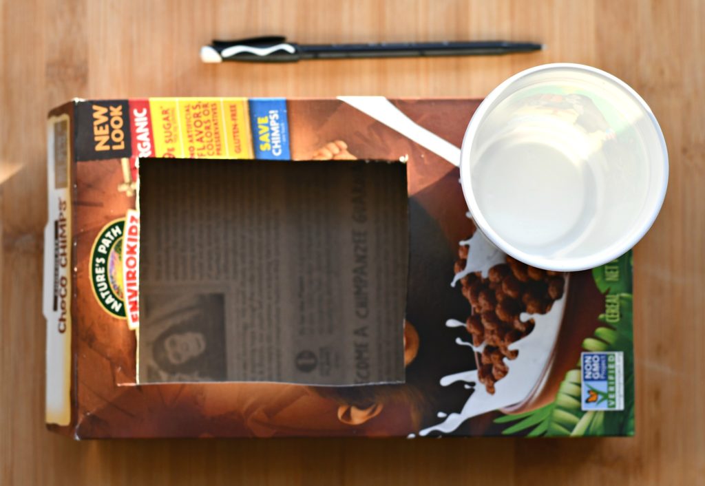 An empty cereal box with a rectangle cut out and starting to trace a circle to cut out for a cup