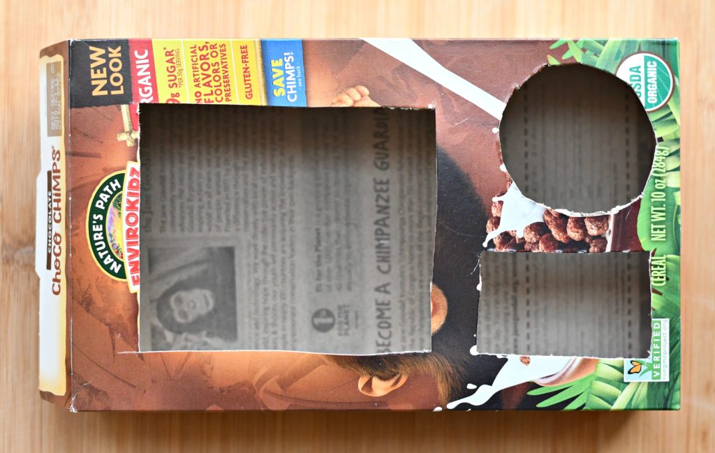 An empty cereal box with a large and small rectangle and circle cut out of it to fit popcorn, a drink and a snack inside.