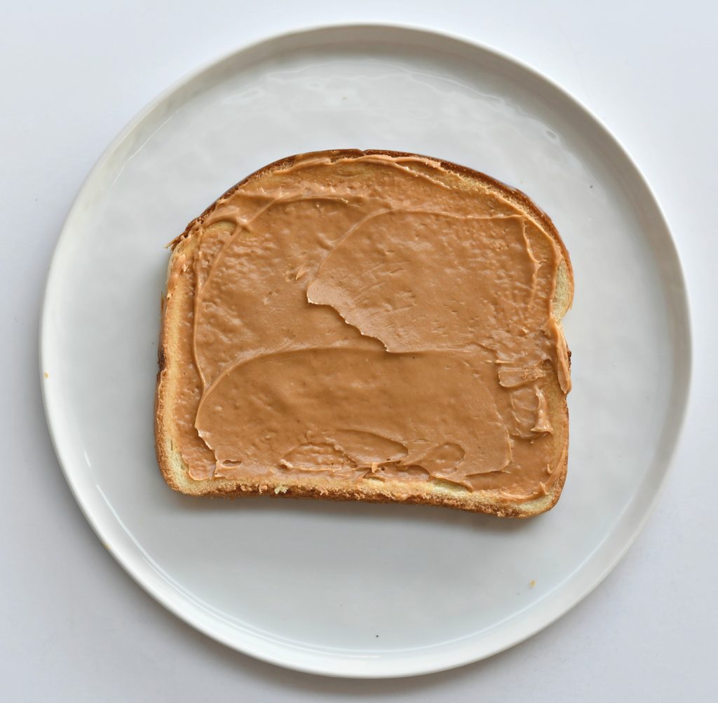 A plate with a piece of bread with peanut butter on it on a white background.