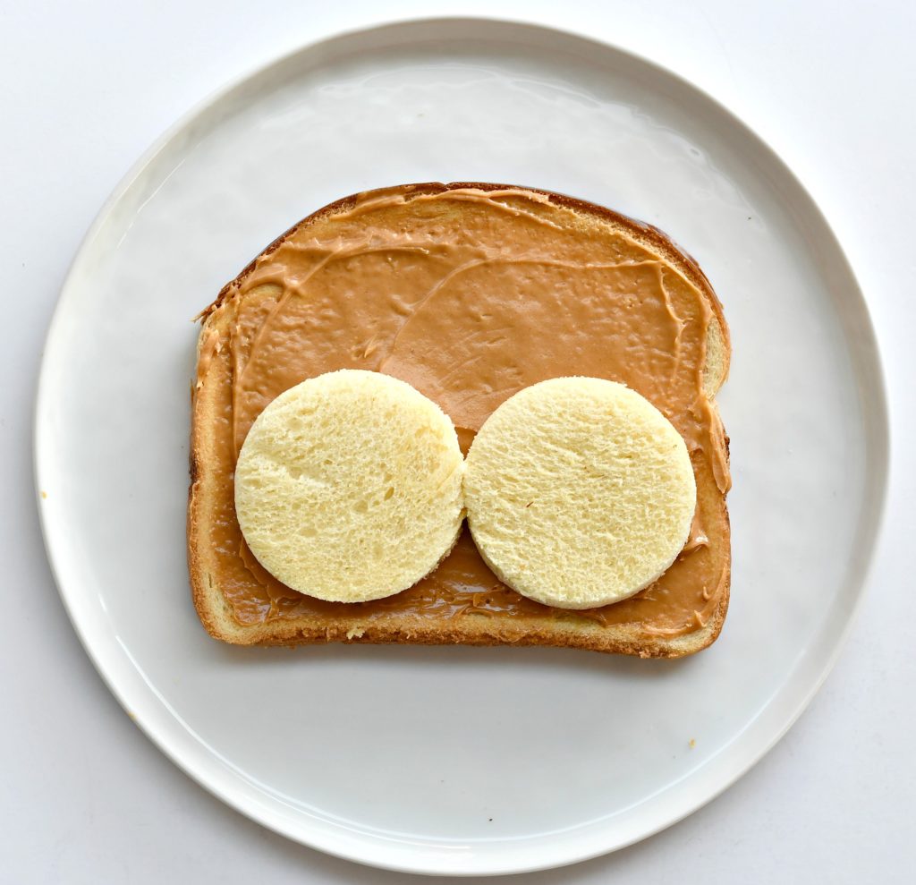 A plate with a piece of toast with peanut butter over it and 2 bread circles for cheeks.