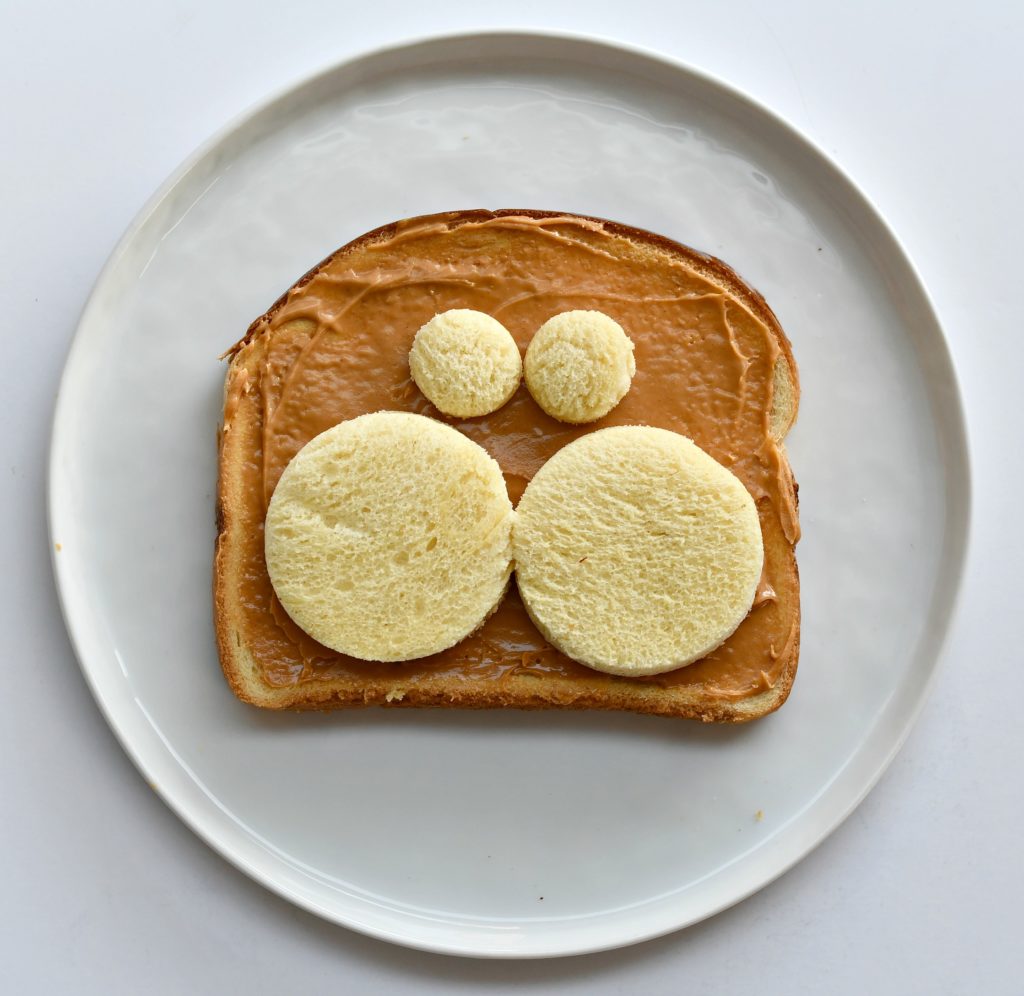 A plate with a piece of toast with peanut butter over it and 2 bread circles for cheeks and 2 for eyes.