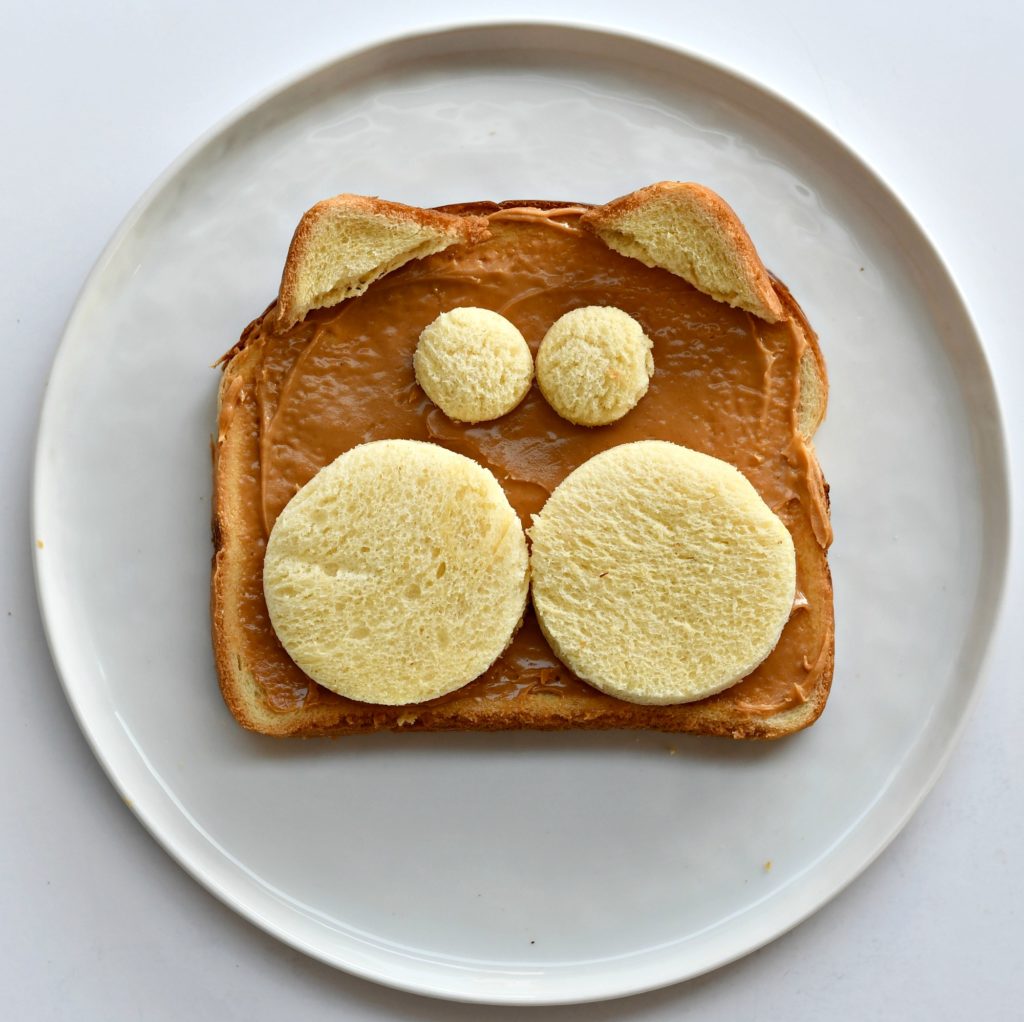 A plate with a piece of toast with peanut butter over it and 2 bread circles for cheeks, 2 for eyes, and 2 corners for ears.