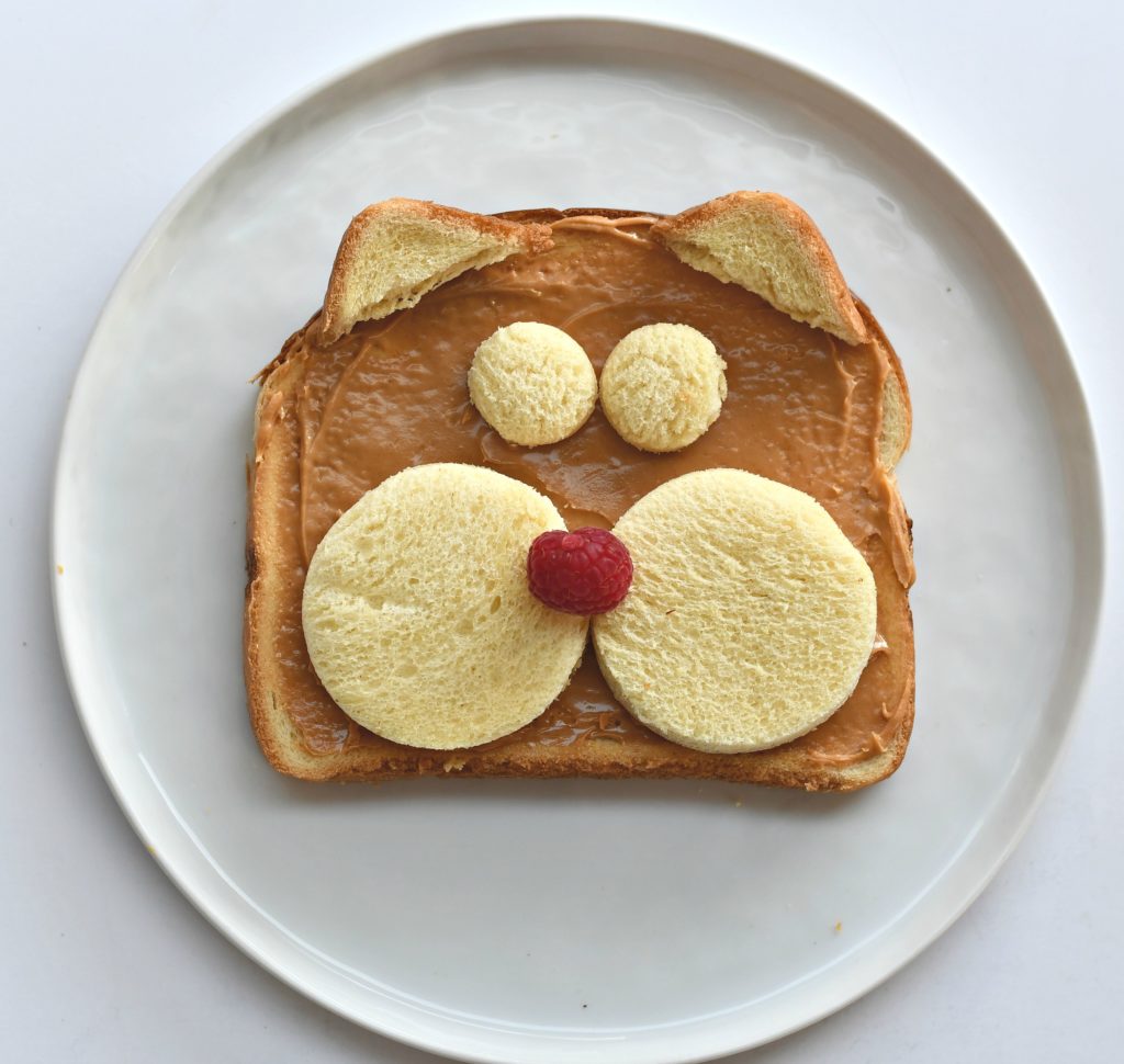 A plate with a piece of toast with peanut butter over it and 2 bread circles for cheeks, 2 for eyes, and 2 corners for ears., and a raspberry nose.