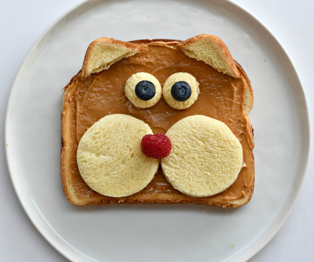 A plate with a piece of toast with peanut butter over it and 2 bread circles for cheeks, 2 for eyes, and 2 corners for ears., a raspberry nose, and blueberry eyes.