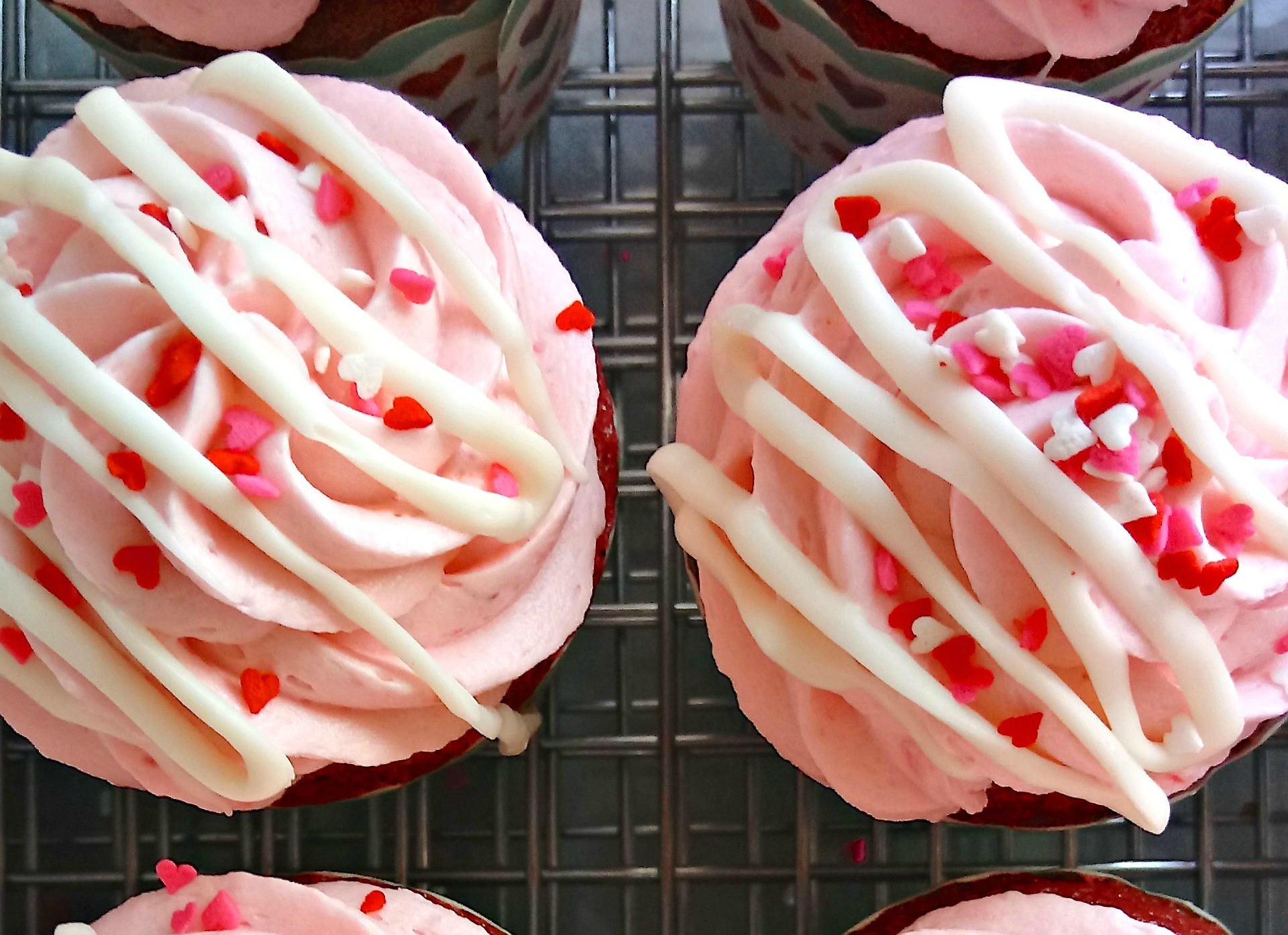 2 Pink Valentines Day cupcakes with a white chocolate drizzle and sprinkles on a wire rack