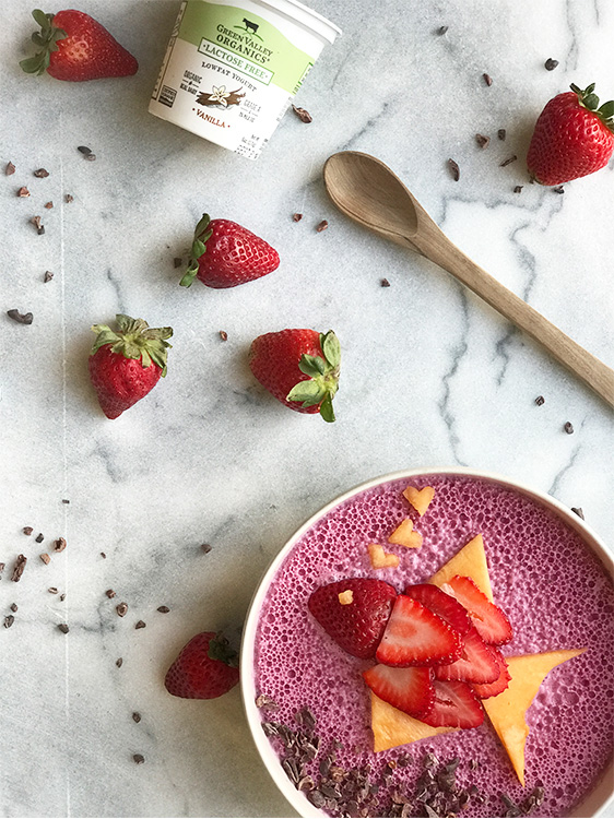 A marble background with a blueberry smoothie bowl and a strawberry fish on top with a hand putting a heart cantaloup on top. 