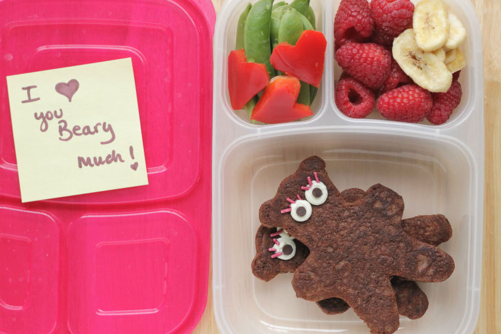 A clear lunchbox container with Teddy Bear-shaped Yogurt Pancakes, heart shaped veggies, and fresh fruit.