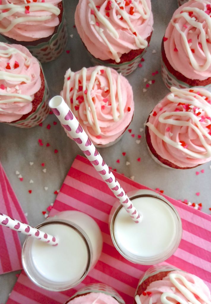 An assortment of Valentines Day Cupcakes with pink frosting and heart sprinkles, served with a couple glasses of milk with straws.