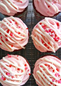A cooling rack with 6 Valentines Day Cupcakes with pink frosting and a white chocolate drizzle.