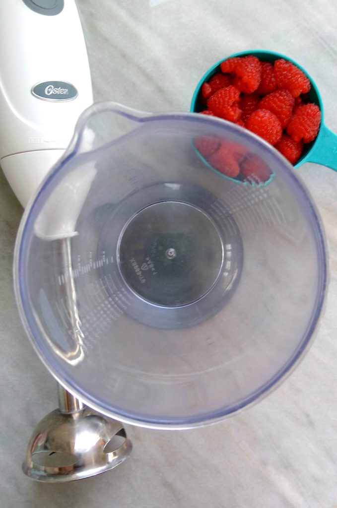 A bowl, immersion blender, and raspberries on a marble background.