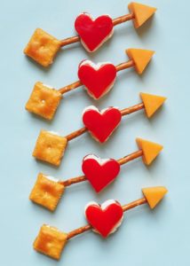 A white background of 5 edible Cupid arrows made with bell peppers, cheese, pretzels, and Cheez-its crackers.