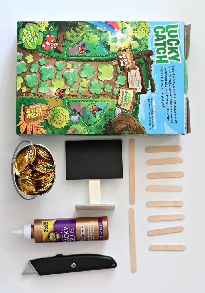 All the supplies needed for a DIY leprechaun trap: a cereal box, pot of gold, glue, a mini chalkboard sign, and popsicle sticks.