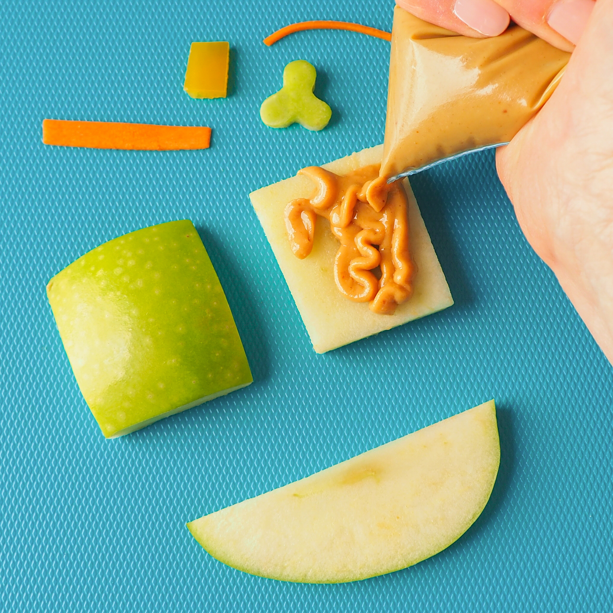 A teal background with cut up apples and a spread of peanut butter for the making of Leprechaun Apple Hats