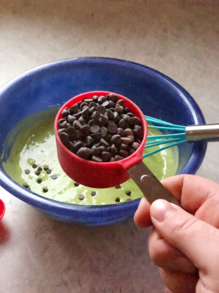A blue bowl filled with mint chocolate chip pancakes batter and a hand adding chocolate chips into it