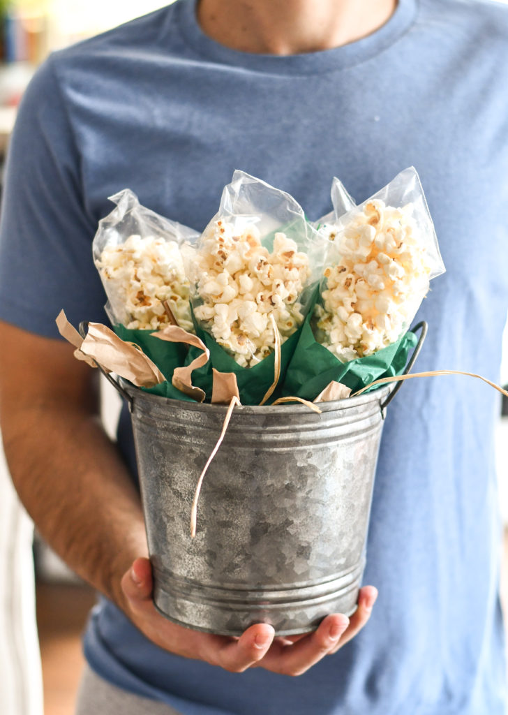 A man's hand holding a bucket full of Popcorn on the Cob Snack Bags