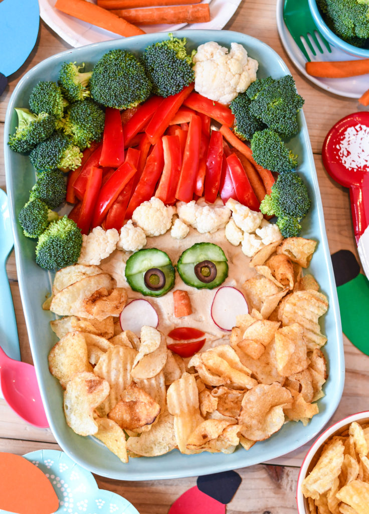 A party table with a santa veggie tray made with veggies and potato chips, along with plates and napkins.