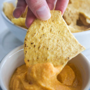 a handing dipping a tortilla chip in a bowl of vegan cheese sauce