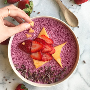 A marble background with a blueberry smoothie bowl and a strawberry fish on top with a hand putting a heart cantaloup on top.