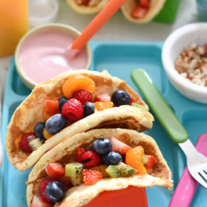 A white table background with rainbow pancake tacos, oj, and silverware.