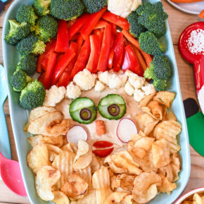 A party table with a santa veggie tray made with veggies and potato chips, along with plates and napkins.